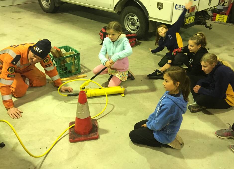One of the Girl Guides uses a hydraulic jack to lift a vehicle, while the rest of the group watch avidly.
